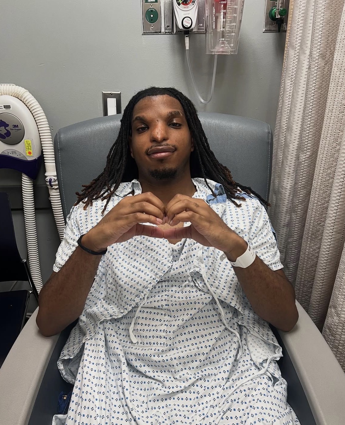 Justin Stewart sitting in a hospital chair, making a heart shape with his hands